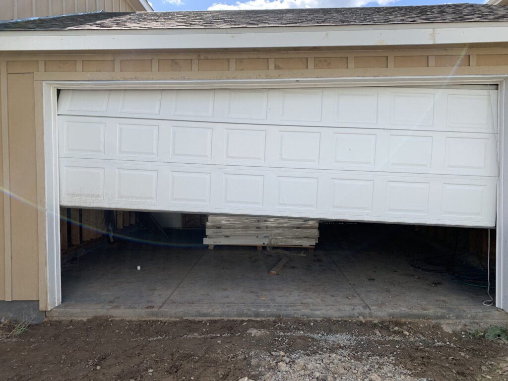 A white garage door is bent. It is on a beige house. The ground is dirt.